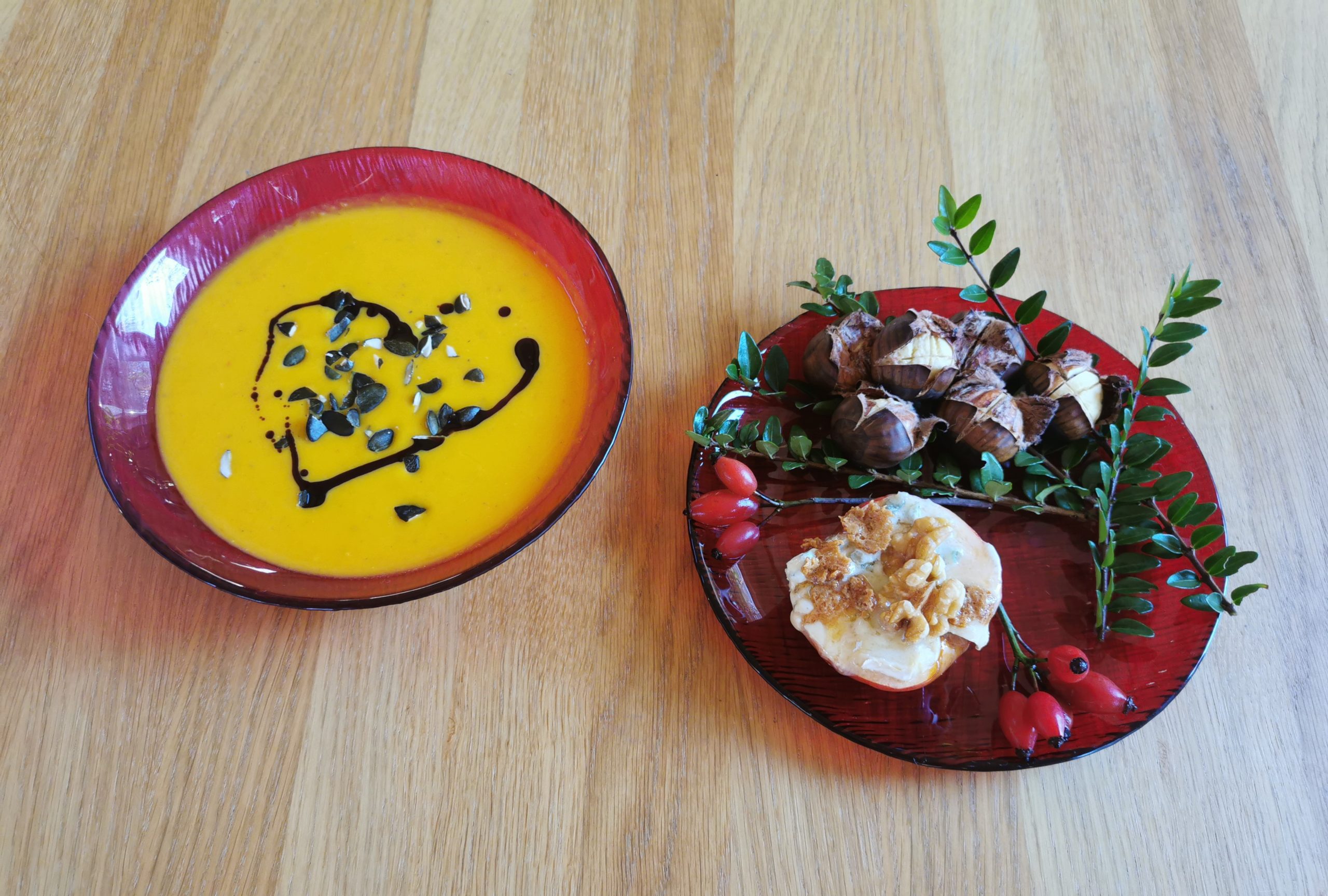 2 rote Teller, links einer mit Kürbiscremesuppe mit gehackten Kürbiskernen und Kernöl, rechts einer mit Maroni und einem gebratenen Apfel mit Gorgonzola und Walnüssen