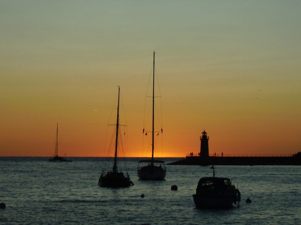 3 Segelboote liegen in der Abendstimmung im Hafen; am Horizont, hinter einem Leuchtturm, verschwindet die orange-gelbe Sonne im Meer