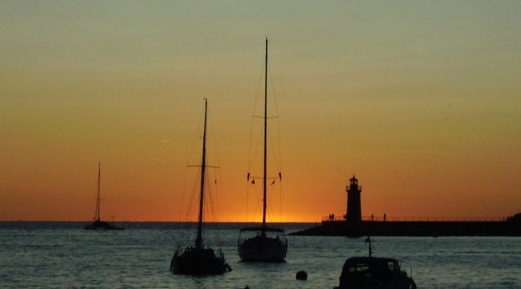 3 Segelboote liegen in der Abendstimmung im Hafen; am Horizont, hinter einem Leuchtturm, verschwindet die orange-gelbe Sonne im Meer